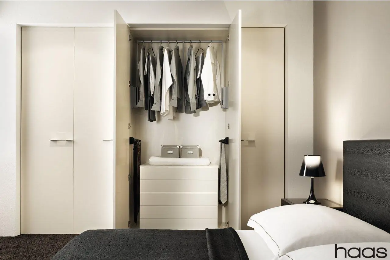 Geöffneter Kleiderschrank mit Hemden auf der Stange, Kommode und ausgeschwenkten Krawattenhaltern