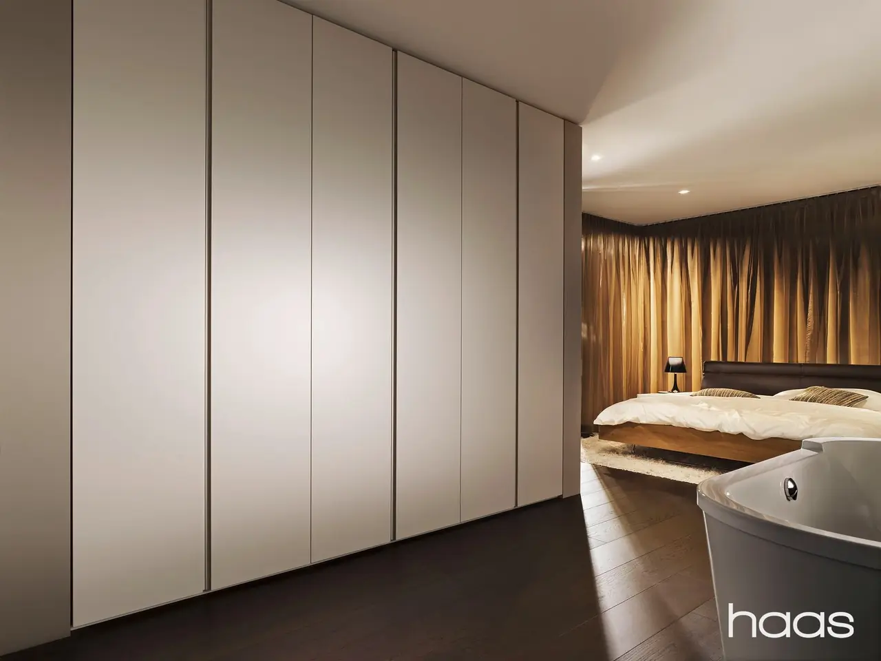 Schlafzimmer mit grifflosen Schrankfronten in Creme, dahinter ein Bett und eine freistehende Badewanne
